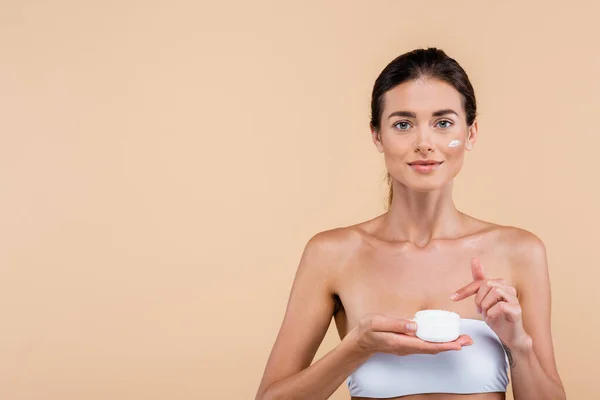 Vista frontal de la mujer joven apuntando al recipiente de crema cosmética aislado en beige - foto de stock