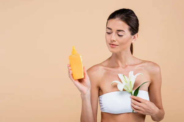 Mujer joven posando con flor de lirio blanco y protector solar aislado en beige - foto de stock