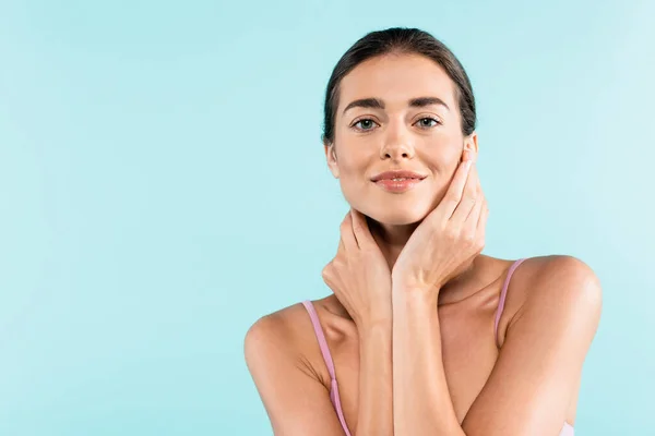 Femme souriante regardant la caméra tout en posant avec les mains sur le cou isolé sur bleu, concept de beauté — Photo de stock