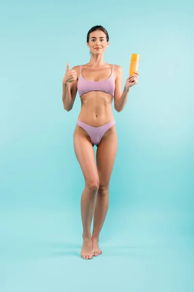 Happy barefoot woman in swimsuit showing thumb up and sunscreen on blue — Fotografia de Stock