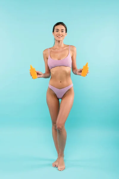 Alegre mujer descalza en traje de baño posando con botellas de bloqueador solar naranja sobre azul - foto de stock