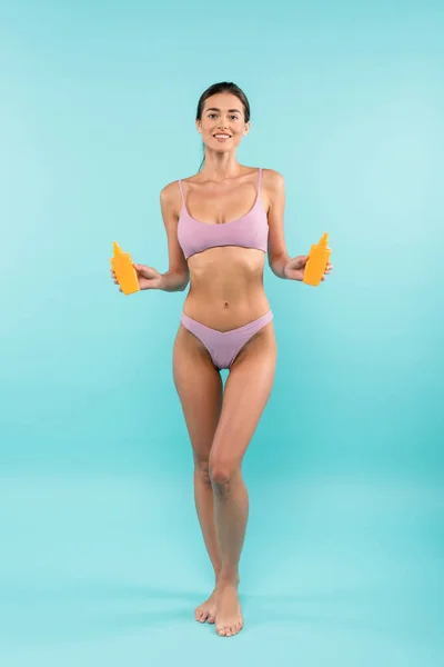 Cheerful barefoot woman in swimsuit posing with orange bottles of sunblock on blue — Stock Photo