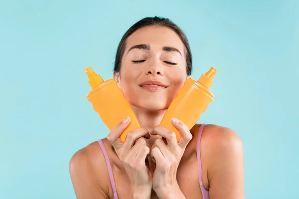 Pleased woman with spray bottles of sunscreen posing with closed eyes isolated on blue — Stock Photo