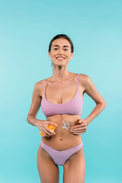 Joyful woman in swimsuit applying sunscreen isolated on blue — Fotografia de Stock