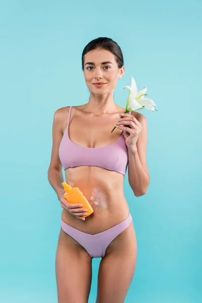 Slim woman with lily flower and sunblock looking at camera isolated on blue — Stock Photo