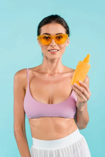 Femme heureuse en maillot de bain et lunettes de soleil orange tenant crème solaire isolé sur bleu — Photo de stock