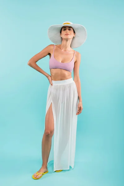 Jeune femme en maillot de bain, paréo blanc et chapeau de soleil posant main sur hanche sur bleu, concept beauté — Photo de stock