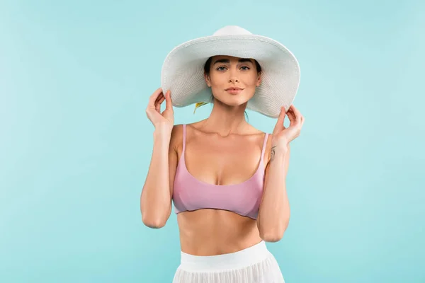 Чарівна жінка в купальнику і парео, торкаючись сонячного капелюха ізольована на синьому, концепція краси — стокове фото