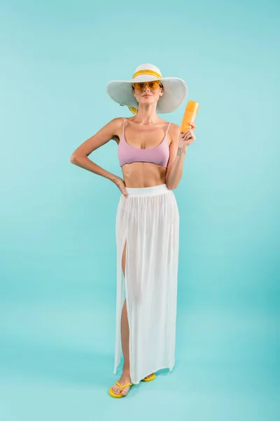 Vue pleine longueur de la femme en chapeau de soleil et paréo blanc posant avec écran solaire sur bleu — Photo de stock