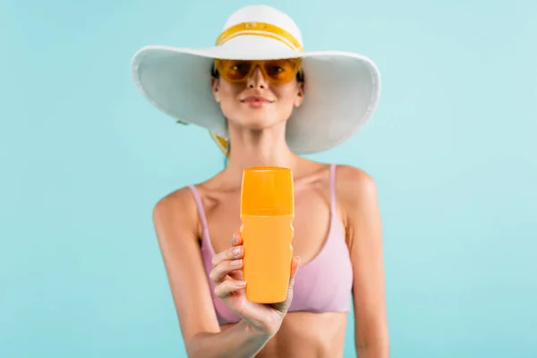 Femme floue en chapeau de soleil et lunettes de soleil orange montrant bouteille de crème solaire isolé sur bleu — Photo de stock