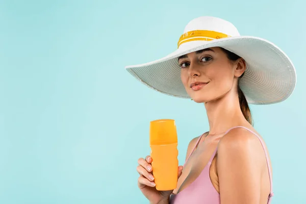 Bonita mujer en sombrero de sol sosteniendo botella de protector solar aislado en azul - foto de stock