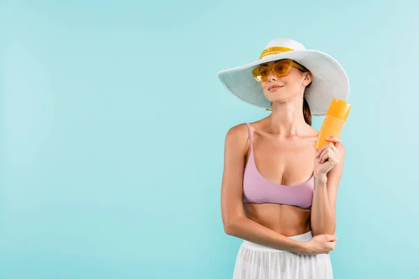 Усміхнена жінка в сонячному капелюсі і парео позує з сонцезахисним кремом ізольовано на синьому — стокове фото
