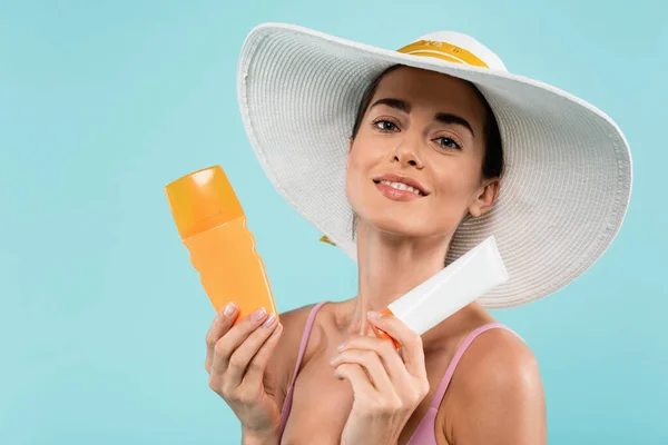 Радісна жінка в сонячному капелюсі позує з трубкою і пляшкою сонячного блоку ізольована на синьому — стокове фото