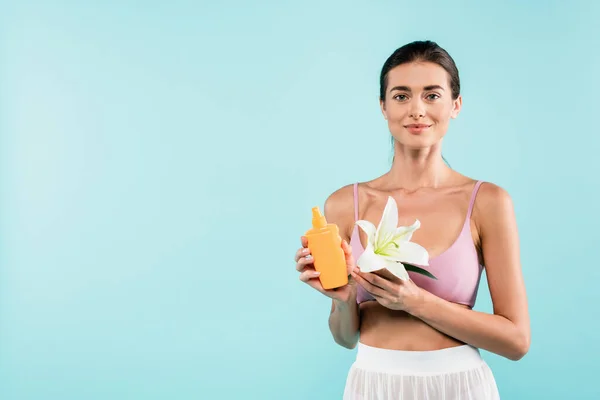 Young woman with bottle of sunscreen and white lily looking at camera isolated on blue — Fotografia de Stock