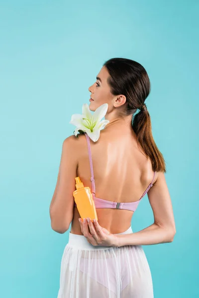 Back view of brunette woman posing with white lily and sunscreen isolated on blue — Fotografia de Stock