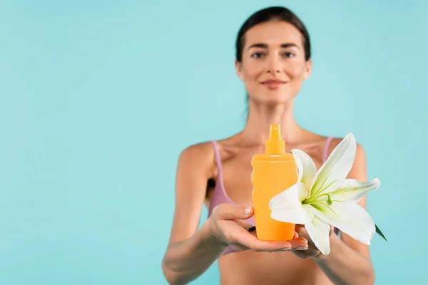 Mujer borrosa sosteniendo lirio blanco y botella de protector solar aislado en azul - foto de stock