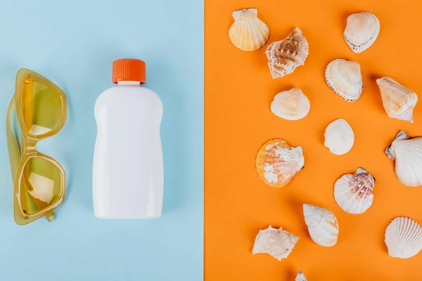 Vista superior de la botella de protector solar blanco cerca de conchas marinas sobre fondo azul y naranja - foto de stock