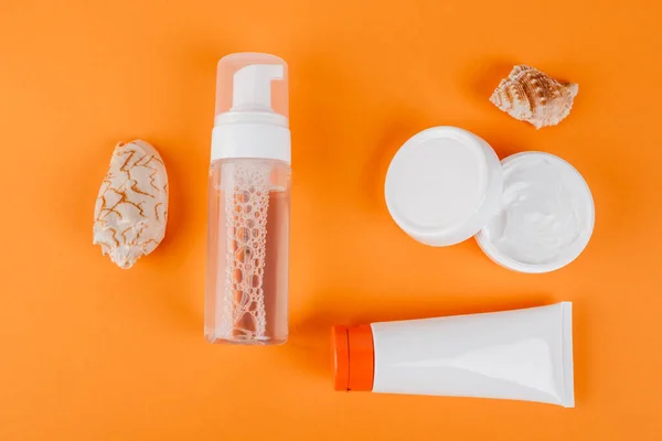 Top view of containers with cosmetic cream, face tonic and sunscreen near seashells on orange — Stock Photo