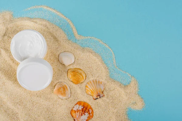 Muscheln in der Nähe kosmetischer Creme auf Sand und blauer Oberfläche — Stockfoto