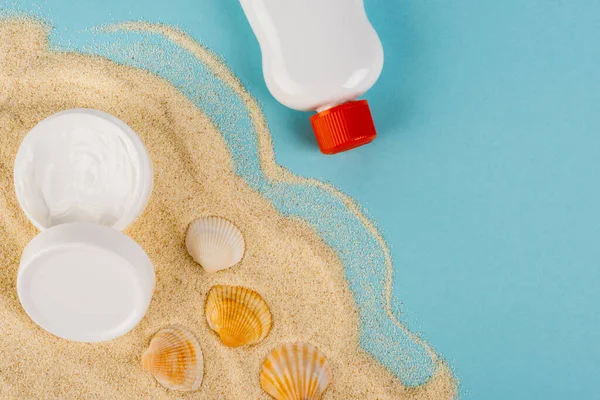 Sonnencreme und kosmetische Creme in der Nähe von Muscheln auf Sand und blauer Oberfläche — Stockfoto