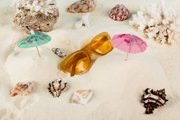 Vista dall'alto di occhiali da sole arancioni vicino a conchiglie e ombrelloni da cocktail sulla sabbia — Foto stock