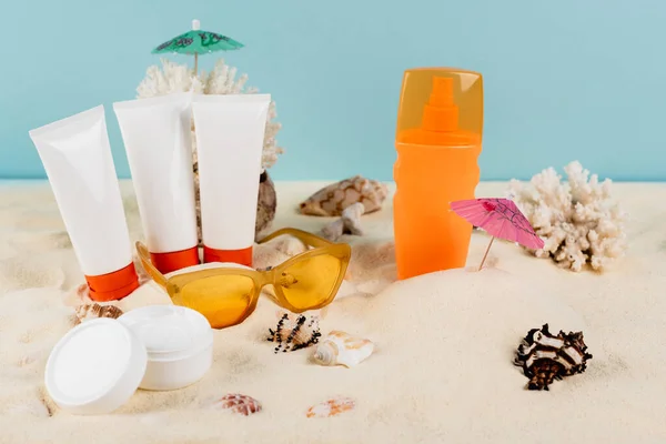 Cosmetic cream, sunblock tubes, seashells and sunglasses on sand isolated on blue — Stock Photo