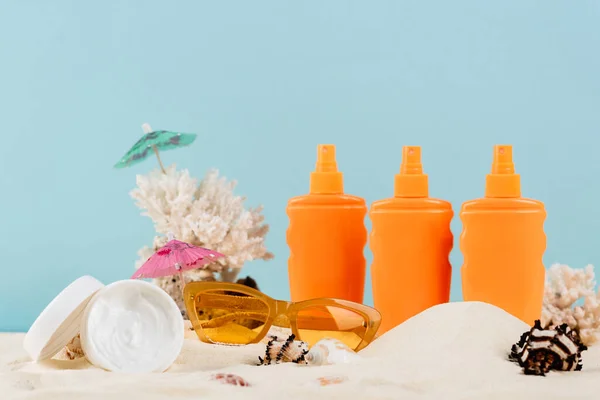 Behälter mit Sonnencreme und Kosmetikcreme in der Nähe von Sonnenbrillen auf Sand isoliert auf blau — Stockfoto