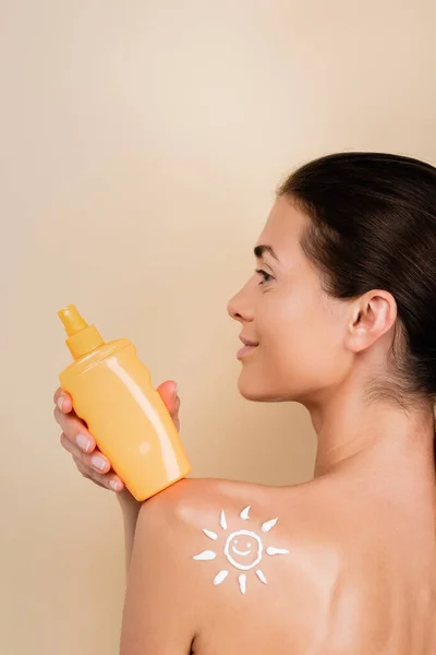 Femme souriante avec l'image du soleil souriant sur l'épaule tenant crème solaire isolé sur beige — Photo de stock
