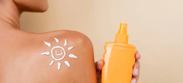 Partial view of woman with bottle of sunblock and image of happy sun on naked shoulder isolated on beige, banner — Stock Photo