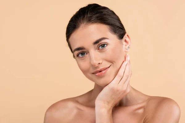 Bonita mujer con hombros desnudos posando con la mano cerca de la cara aislada en beige, concepto de belleza - foto de stock