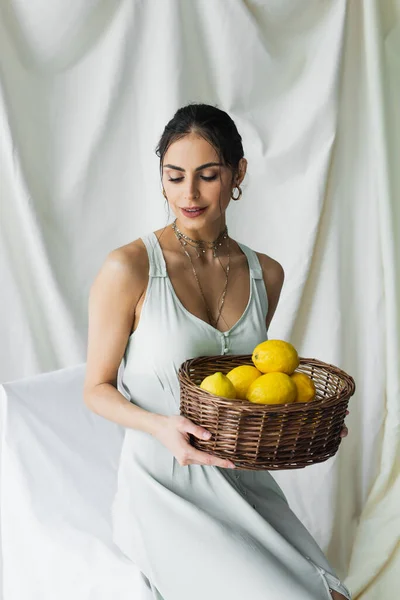 Happy woman in dress holding wicker basket with lemons on white — Stock Photo