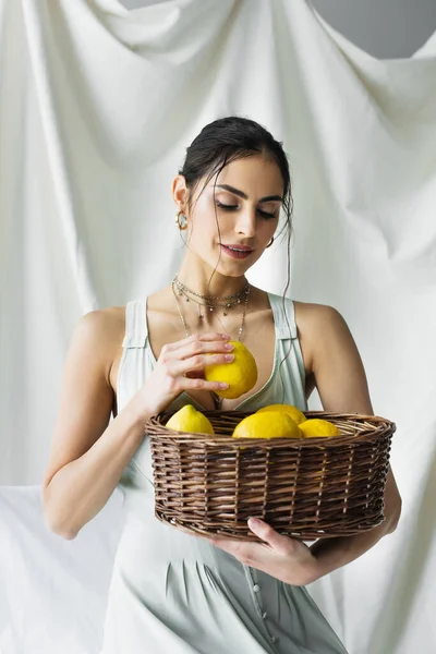 Femme souriante en robe tenant panier en osier avec des citrons sur blanc — Photo de stock