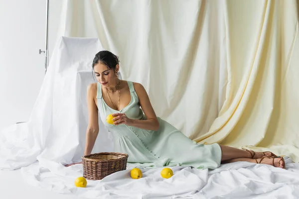 Pleine longueur de jolie femme en robe tenant citron et regardant panier en osier sur blanc — Photo de stock