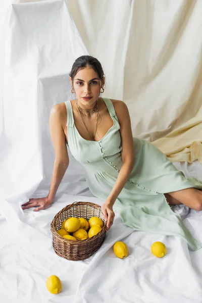 Vista de ángulo alto de la mujer bonita en vestido sentado cerca de la canasta de mimbre con limones en blanco - foto de stock