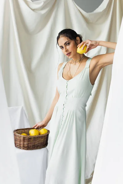 Pretty woman in dress holding ripe lemon near wicker basket on white — Stock Photo