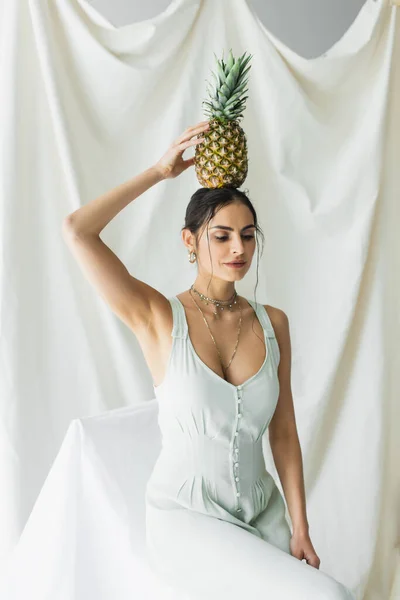 Brunette woman in dress holding pineapple above head on white — Stock Photo