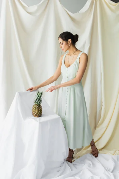 Full length of brunette woman in dress posing near ripe pineapple on white — Stock Photo