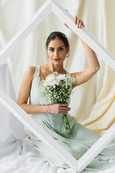 Brunette woman holding bouquet of blooming flowers and posing near frame on white — Stock Photo