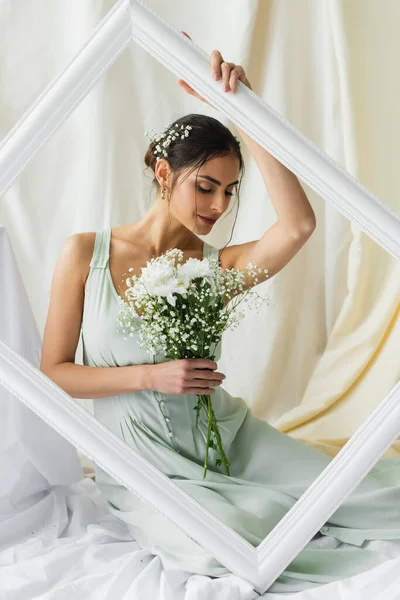 Donna sorridente che tiene mazzo di fiori in fiore e posa vicino cornice su bianco — Foto stock