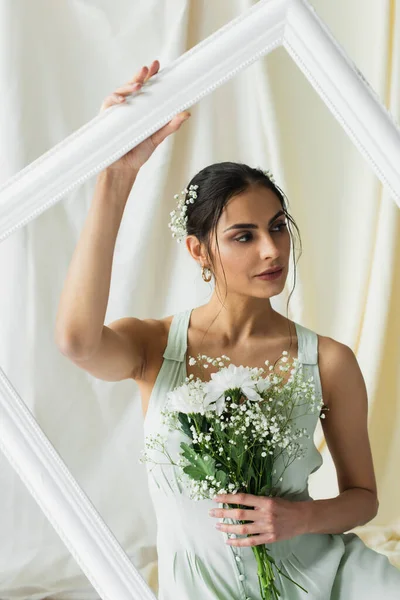 Brünette Frau, die einen Strauß blühender Blumen in der Hand hält und in der Nähe eines Rahmens auf weißem Grund wegsieht — Stockfoto