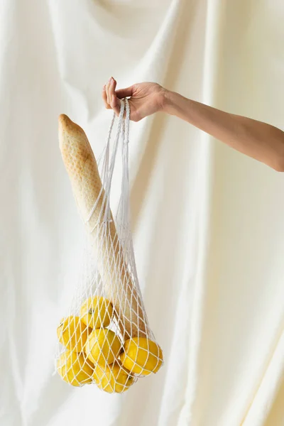 Vista cortada da mulher segurando saco de corda reutilizável com limões e baguete no branco — Fotografia de Stock