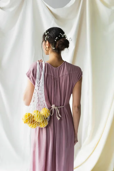 Back view of brunette woman holding reusable string bag with lemons on white — Stock Photo