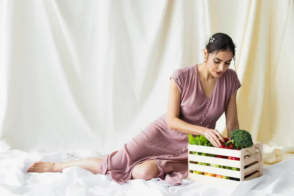 Brunette woman reaching red bell pepper near vegetables in wooden container on white — Stock Photo