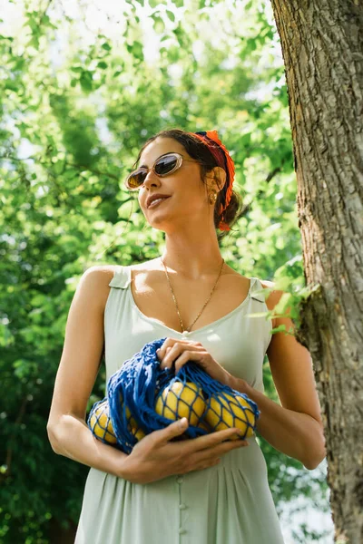 Mulher feliz em óculos de sol e vestido segurando saco de corda com limões — Fotografia de Stock