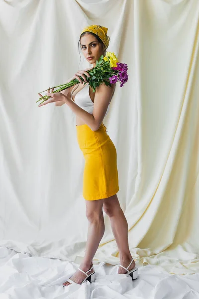 Pleine longueur de jolie femme en foulard jaune tenant des fleurs sur blanc — Photo de stock