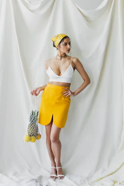 Mulher em lenço amarelo e top de colheita posando com saco de cordas cheio de limões e abacaxi no branco — Fotografia de Stock