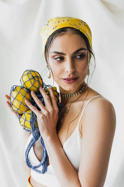 Woman in yellow headscarf and crop top holding string bag with lemons on white — Stock Photo