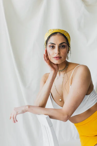 Femme à la mode en foulard jaune et haut de culture assis sur chaise sur blanc — Photo de stock