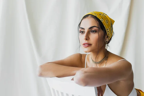 Mulher na moda em lenço de cabeça amarelo e top de cultura encostado na cadeira no branco — Fotografia de Stock