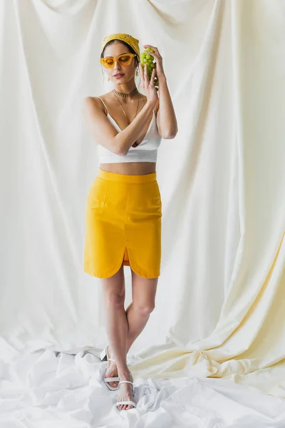 Pleine longueur de femme à la mode en lunettes de soleil jaunes, foulard et crop top exploitation raisins sur blanc — Photo de stock